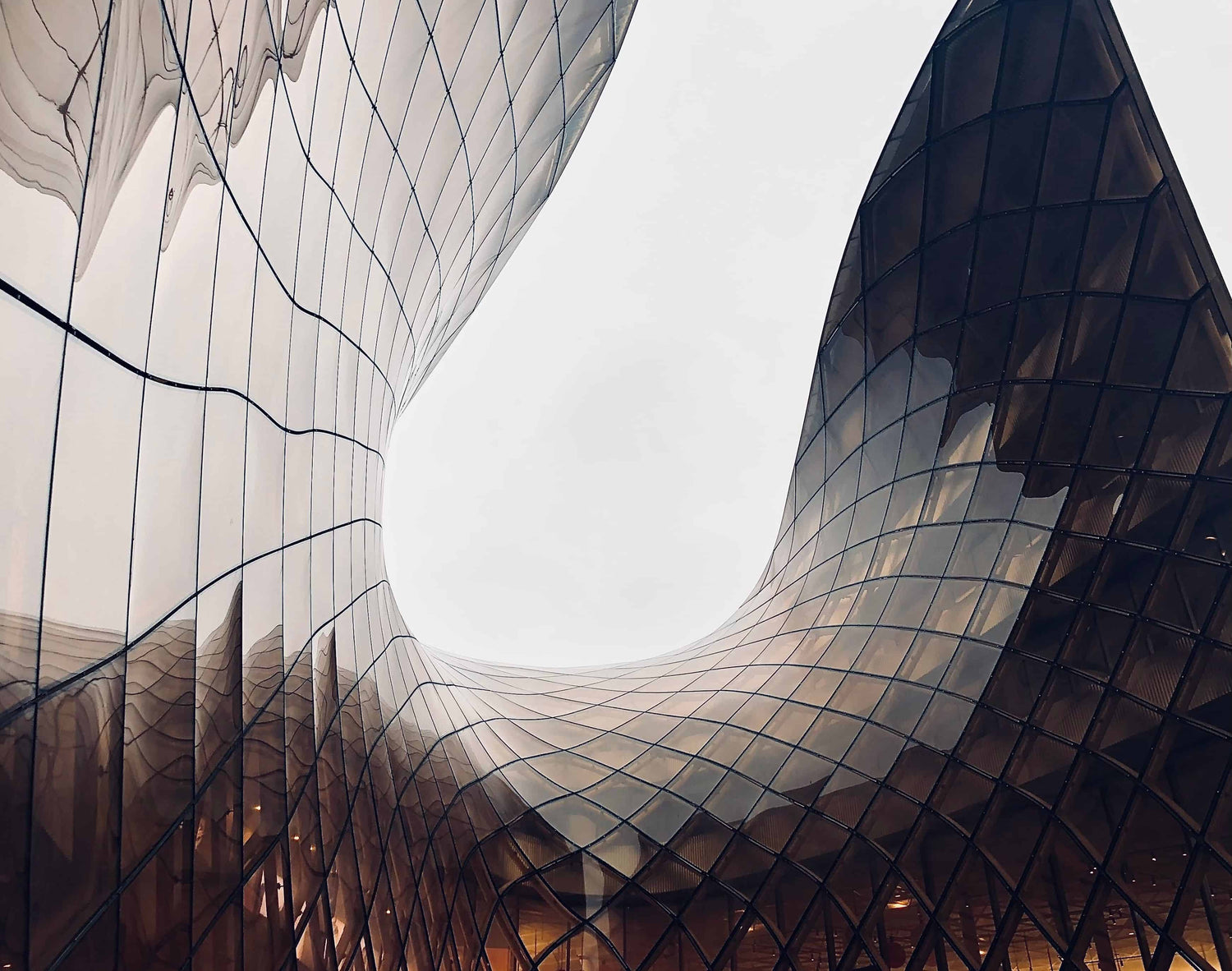 Modern architectural structure with curved glass walls and a reflective grid facade, photographed from below against an overcast sky.