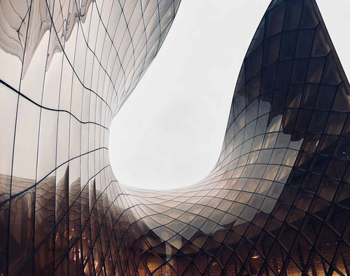 Modern architectural structure with curved glass walls and a reflective grid facade, photographed from below against an overcast sky.