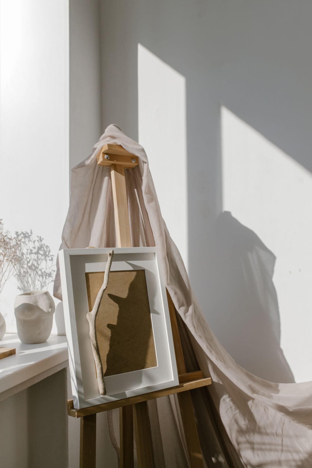 A wooden easel in a sunlit room holds a white-framed artwork draped with sheer beige fabric. Strong window shadows fall across a white wall and windowsill, with a small vase of dried stems on the left.