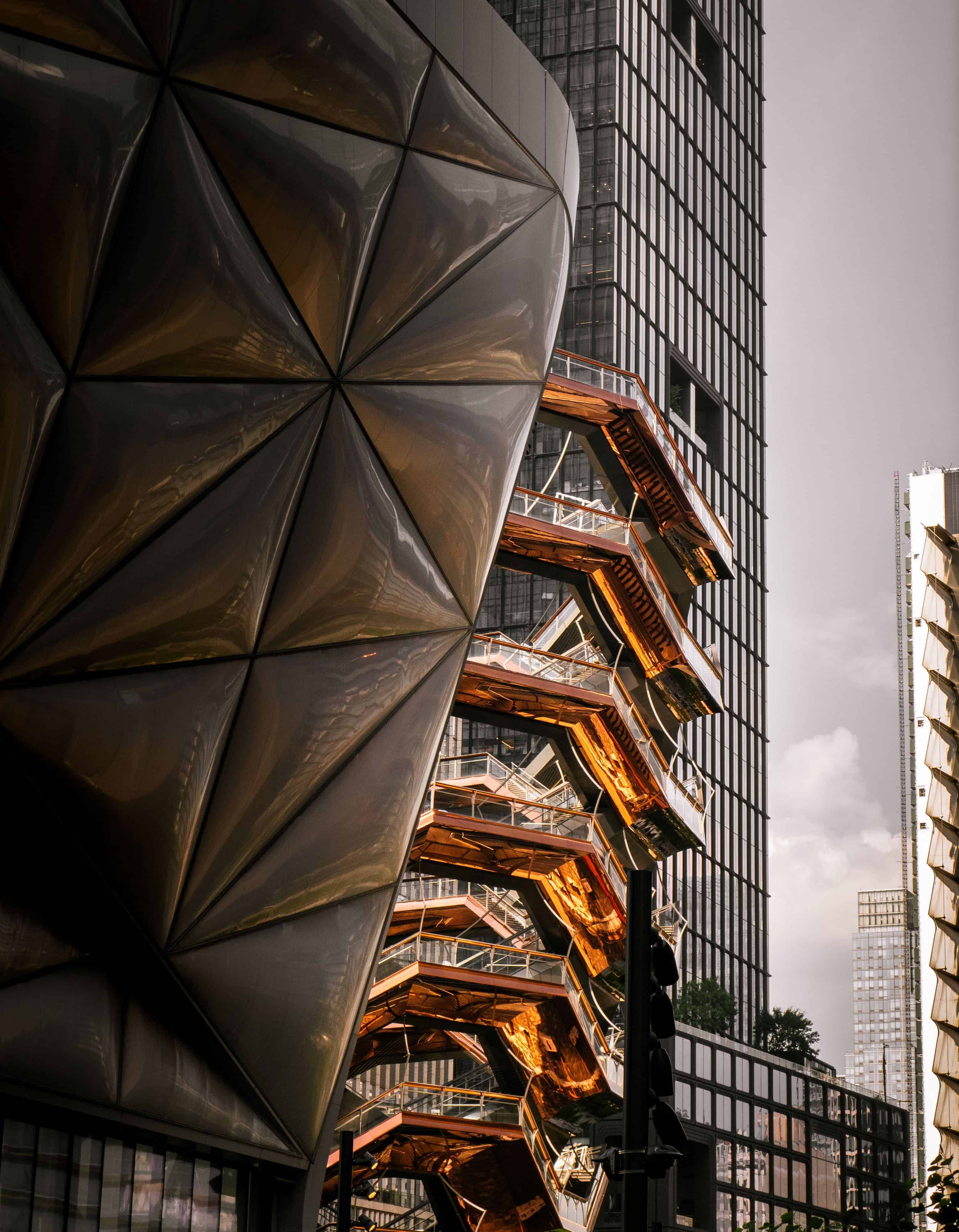 Low-angle photograph of the Vessel on the Walk in New York City, showcasing its copper-colored geometric steel structure against the dark facade of the nearby Comcast Center building.