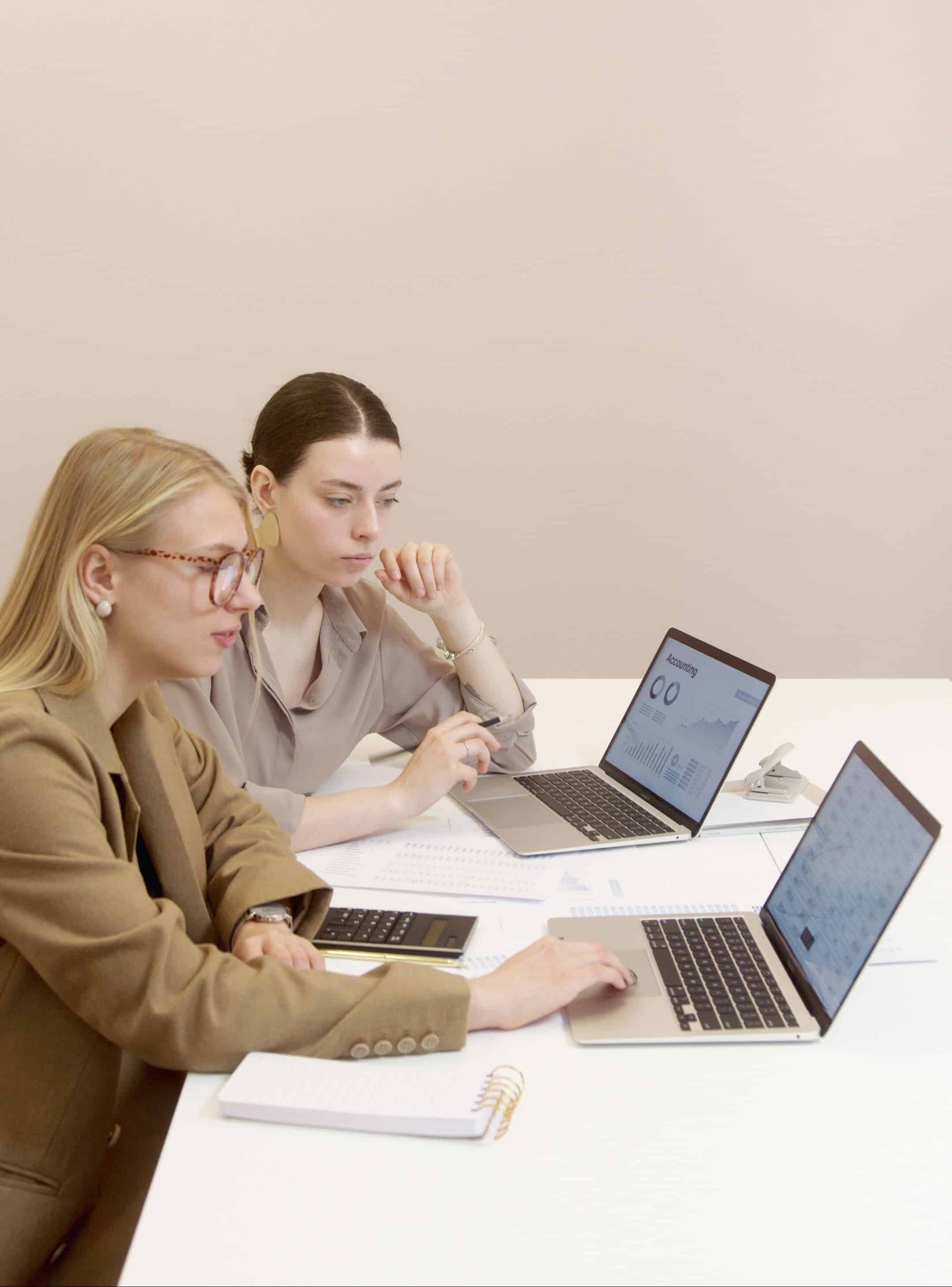 Two women collaborating at a white table with laptops in a bright, modern office setting.