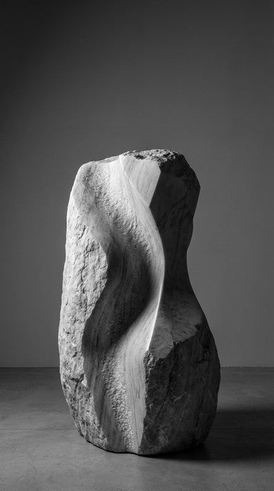 A large abstract stone sculpture with organic, flowing curves and rough textured surfaces, photographed in a minimalist studio setting with a dark gray background.