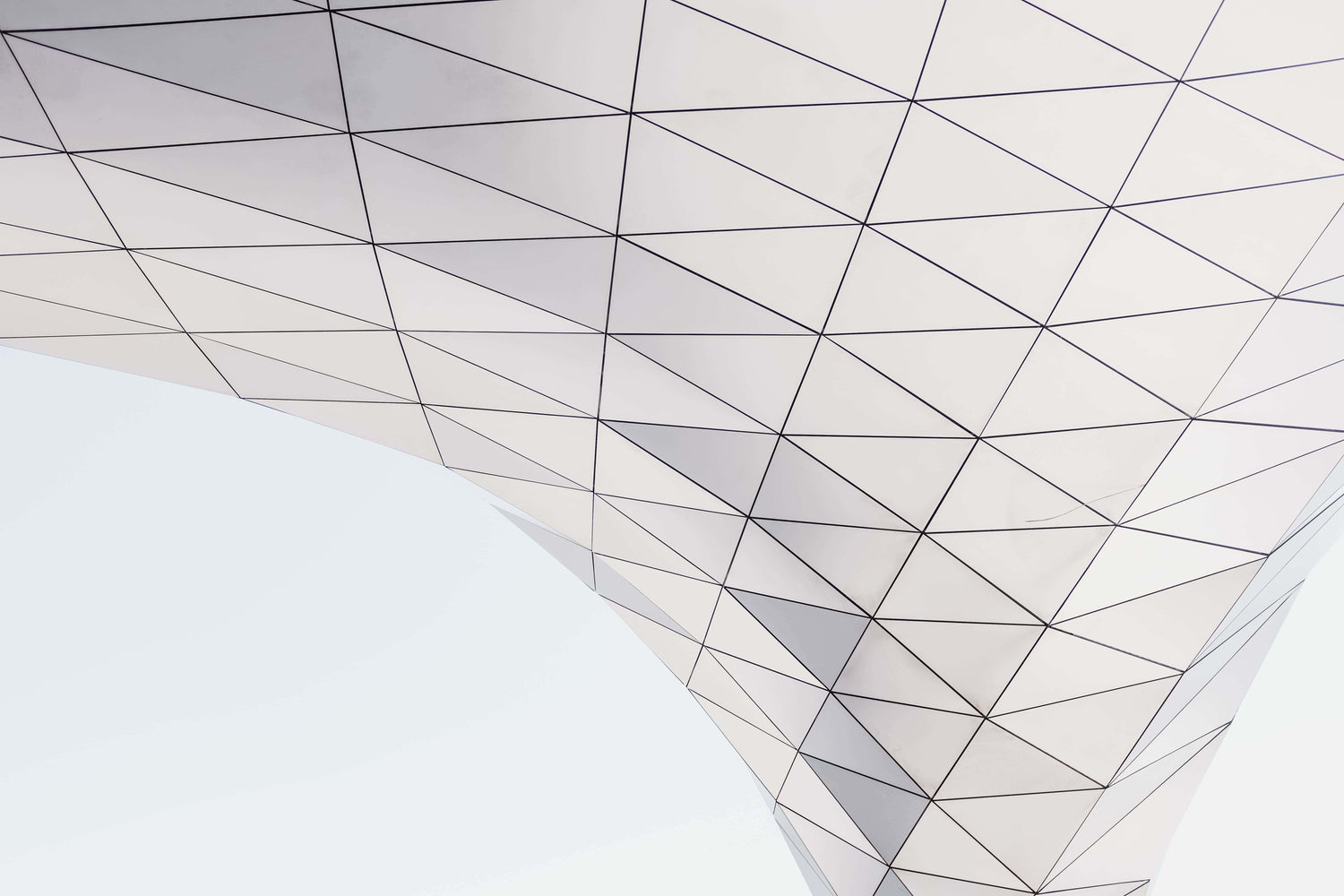 Abstract architectural photograph of a white and gray geometric structure with a triangular lattice pattern, viewed from below against a pale sky.